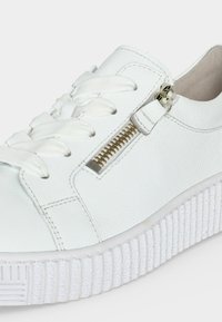 Baskets en cuir blanc avec semelle épaisse texturée, lacets blancs et détail de fermeture éclair dorée sur le côté, sur un fond uni.