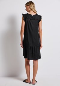 Schwarzes Kleid aus weichem Stoff, mit kurzen Rüschärmeln und einem gestuften Design. Auffälliges Detail: zarte Spitzenborte am Rücken.