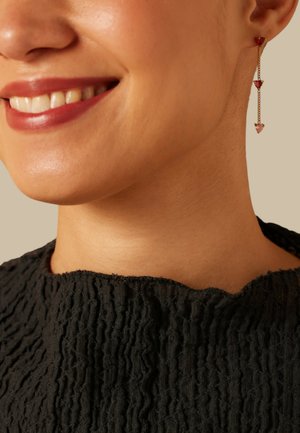 Oorbellen met goudkleurige hangers met drie hartvormige edelstenen in verloopkleuren van rood en roze. Top van gestructureerde zwarte stof met een hoge halslijn.