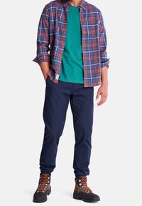 Koszula w kratę button-down w kolorze czerwonym, niebieskim i białym na tle niebieskiej koszulki; granatowe zwężane spodnie i brązowe buty trekkingowe z pomarańczowymi sznurówkami.