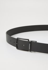 Μαύρη υφασμάτινη ζώνη από δέρμα με ορθογώνια μαύρη μεταλλική πόρπη. Έχει λεπτομέρεια λογότυπου και πολλές τρύπες ρύθμισης κατά μήκος της ζώνης.