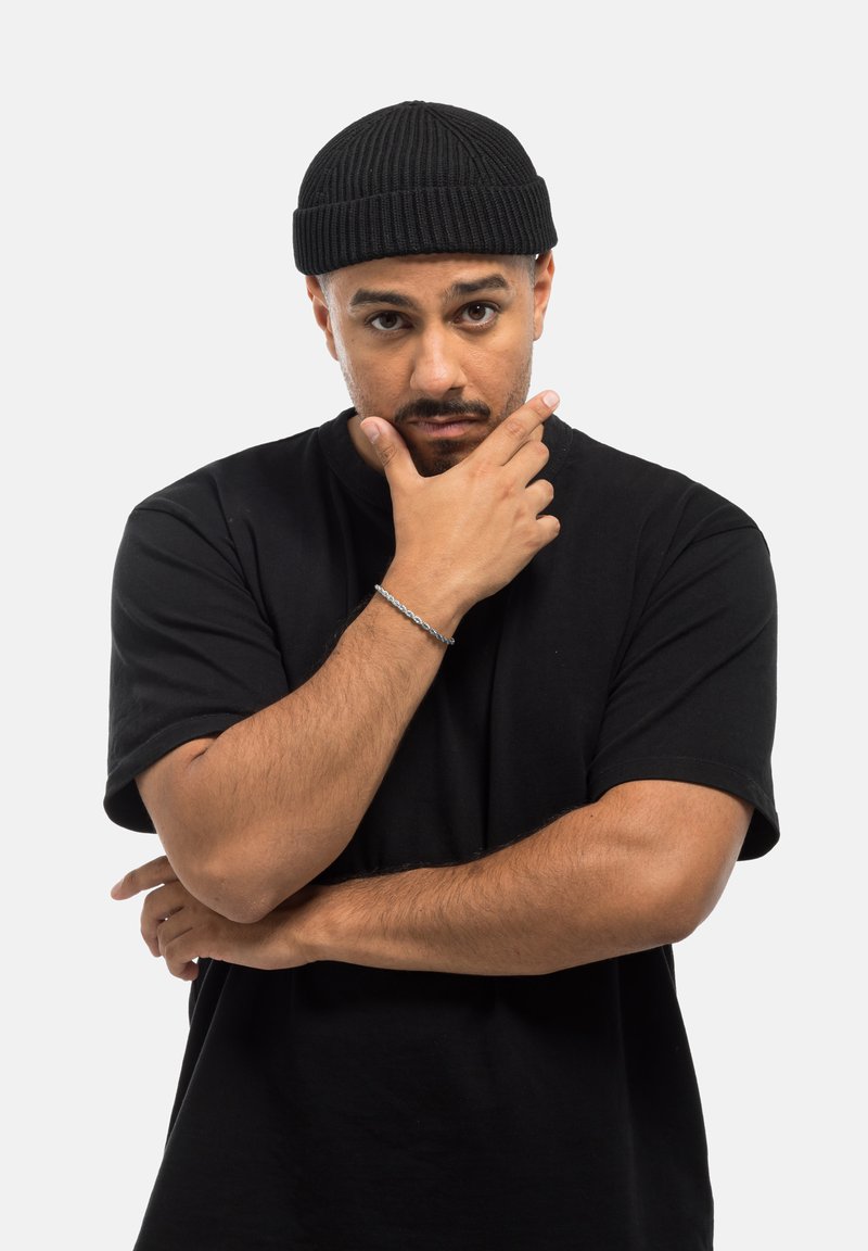 Bonnet en tricot noir posé sur un t-shirt noir à manches courtes. La personne a une expression neutre, avec une main reposant sur le menton. Détail de bracelet en argent.