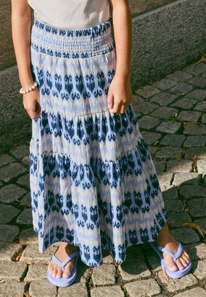 Enfant portant une jupe à volants bleue et blanche à motifs, un haut blanc, un bracelet de perles et des tongs violet clair, debout sur un pavé en pierres.