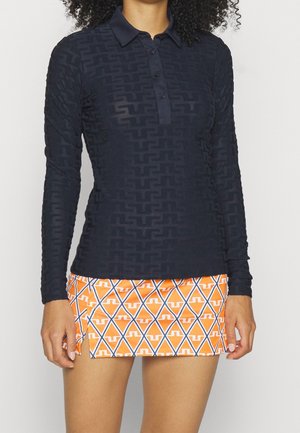 Kobieta nosząca dopasowaną, granatową koszulkę polo z długim rękawem z subtelnym geometrycznym wzorem oraz krótką pomarańczową spódnicę z niebieskimi i białymi rombami.