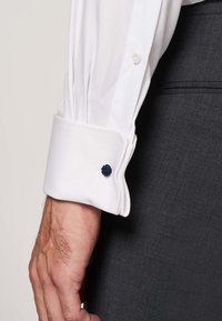 Poignet de chemise blanche présentant une texture unie, avec un accent de bouton décoratif bleu. Main montrant un veston gris.
