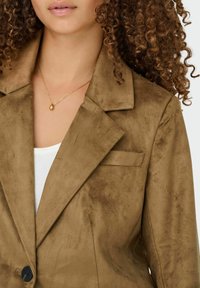 Bruine suède blazer met gekartelde revers, enkele knoopsluiting en een borstzak. Gelaagd over een witte top. Gouden druppelvormige ketting.