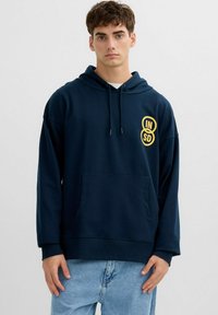 Joven con una sudadera con capucha azul marino con el logo de círculos amarillos "IN SD" en el pecho, jeans azul claro, de pie contra un fondo liso y claro.
