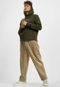 Olivengrønn hettegenser med frontlomme og snører, sammen med lysebrune kordfløyelsbukser. Modellen har på seg grå sneakers.