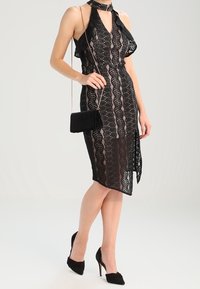 Robe en dentelle noire avec un décolleté halter, présentant un design fluide et des motifs complexes. Associée à un petit sac à main noir et des talons.