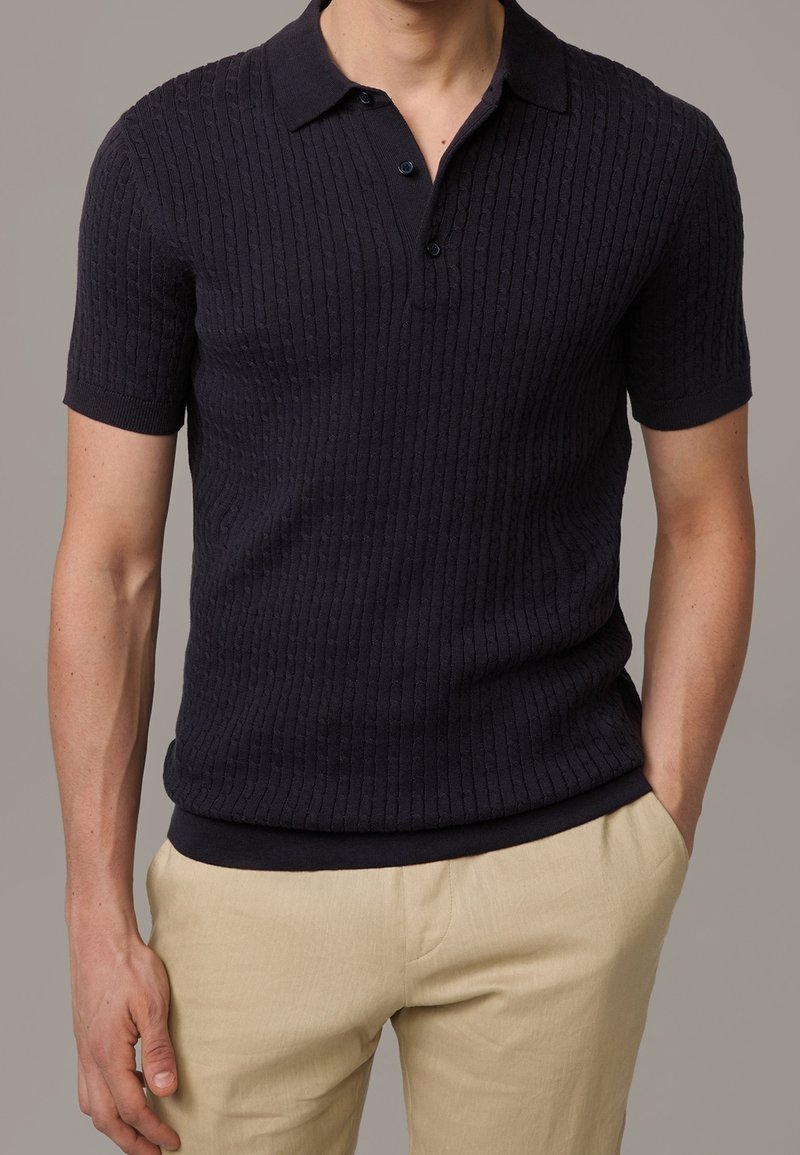 Polo shirt navy à manches courtes avec un design en ribbed texturé, patte de boutonnage à trois boutons et un col uni. Porté avec un pantalon beige.