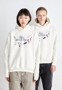 Un sweat à capuche blanc avec un imprimé éclaboussé coloré et le texte "C A L L O O B U C" sur le devant, associé à un pantalon en denim.