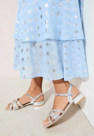 Lichtblauwe doorschijnende rok met zilveren stippen, gecombineerd met zilveren sandalen met lage hak, voorzien van kristallen versieringen en enkelriempjes.