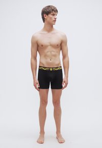 Μαύρα μποξεράκια με διακοσμητική κίτρινη μέση, με ομαλό ύφασμα και εφαρμοστή σχεδίαση, φορεμένα από έναν όρθιο ανδρικό μοντέλο.