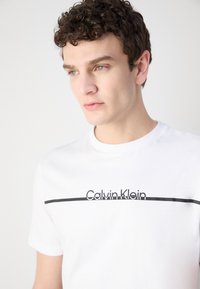 Hvid bomulds T-shirt med et minimalistisk design, der har en sort vandret linje og teksten "Calvin Klein" i kontrasterende sort skrifttype.