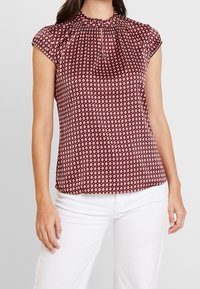 Frau trägt eine kurzärmlige Bluse mit maroon- und weißen Rautenmustern und Schlüssellochausschnitt, kombiniert mit weißen Hosen, vor einem grauen Hintergrund.