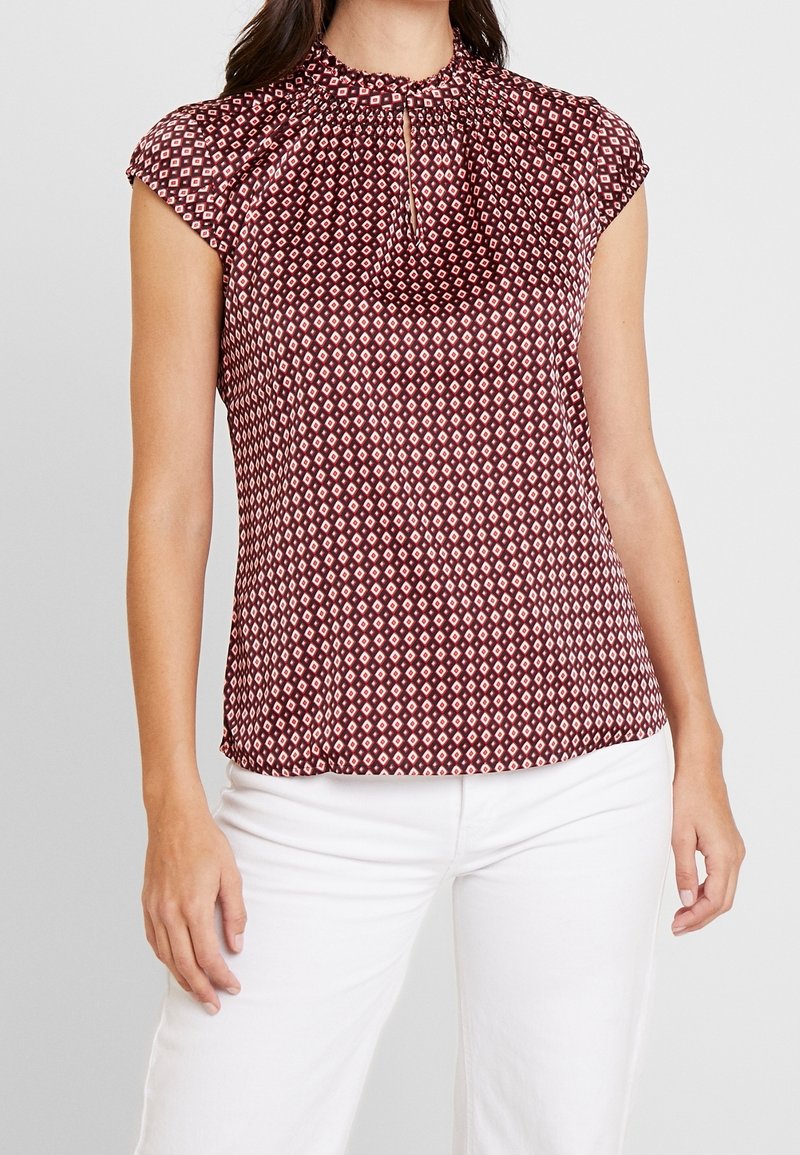 Donna che indossa una blusa a maniche corte a motivo diamante bordeaux e bianco con scollatura a chiave, abbinata a pantaloni bianchi, su sfondo grigio.