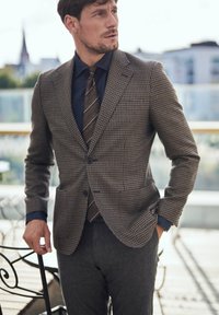 Brązowa marynarka w wzór houndstooth z dwoma przednimi kieszeniami, zestawiona z granatową koszulą i brązowym wąskim krawatem oraz szarymi spodniami o kroju dopasowanym. Teksturowany materiał.