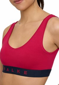 Red sleeveless sports bra with a deep neckline and contrasting navy elastic band featuring "FALKE" logo in maroon. Soft, smooth texture.