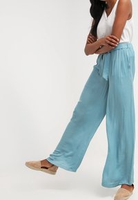 Donna che indossa un pantalone ampio azzurro chiaro, una canotta bianca senza maniche e delle scarpe beige slip-on, in piedi con le braccia incrociate e una gamba leggermente sollevata.
