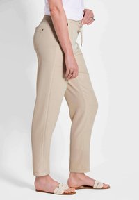 Beige elastische broek met zijzakken, taps toelopende pijpen en een soepele textuur. Gecombineerd met lichtgekleurde platte sandalen.