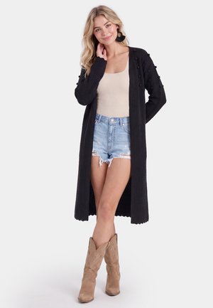 Vrouw die een lange zwarte cardigan draagt, een beige tanktop, gescheurde spijkershorts, bruine cowboylaarzen en grote zwarte oorbellen, staand met één hand bij het gezicht.