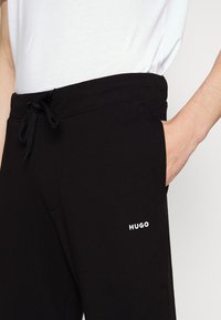 Black sweatpants made of cotton blend, featuring an elastic waistband with a drawstring and side pockets. White "HUGO" logo on leg.