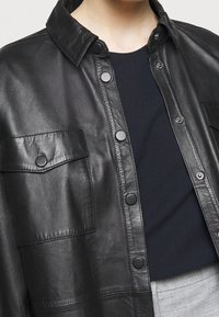 Černá kožená košile bunda s špičatým límečkem, druky, dvěma náprsními kapsami a hladkou texturou, ve spojení s tmavým topem.