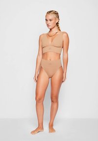 Beige to-delt badetøysett med en bralette-topp med kryssforan-design og høytlivsbukser. Materialet virker glatt og elastisk.