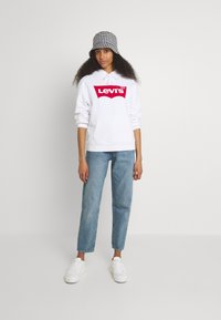 Felpa bianca con logo rosso Levi's, tasca frontale e cordino; abbinata a jeans chiari e sneakers bianche. Cappello a secchiello bianco e nero a scacchi.