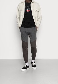 Světlé šedé košilové tričko přes černé tričko s grafikou, doplněné šedými jogger kalhotami s tmavě šedými doplňky a černými teniskami.