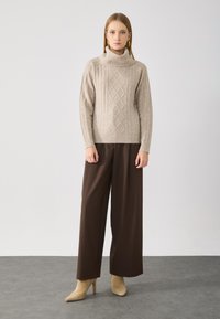 Pull en maille beige à col montant, associé à un pantalon marron ample et des escarpins beiges à bout pointu sur un sol gris.