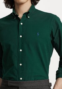 Zielona koszula zapinana na guziki, wykonana z bawełny o teksturowanym wykończeniu, z małym niebieskim logo na lewej piersi oraz białymi guzikami.