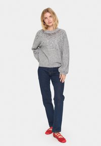 Maglione grigio lavorato a maglia con scollo rotondo, adornato con decorazioni argento e nere. Indossato con jeans blu scuro e scarpe ballerine rosse.