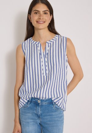 Jeune femme aux longs cheveux foncés portant un chemisier blanc sans manches à rayures verticales bleues et un jean bleu, souriant à la caméra.