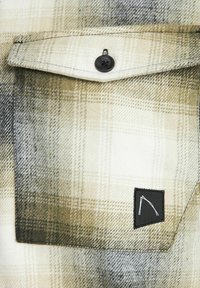Camicia di flanella a quadri con tonalità beige e nere, dotata di una tasca sul petto con un'etichetta nera e dettagli dei bottoni neri. Texture morbida.