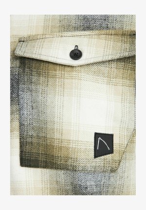 Rutigt flanellskjorta i beige och svart, med en bröstficka som har en svart etikett och svarta knappdetaljer. Mjuk textur.