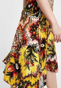 Ljusblommig klänning med gul bakgrund och röda, rosa och gröna mönster. Tillverkad av lättviktig tyg med lagerdrapering.