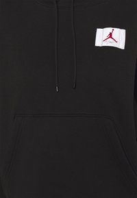 Sweat à capuche noir avec poche avant et cordons, arborant un petit patch blanc avec la silhouette rouge d'un joueur de basket-ball et le texte "Flight" sur la poitrine.