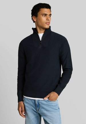 Mann schaut zur Seite, trägt einen dunkelblauen Halb-Reißverschluss-Pullover über einem weißen Hemd und hellblaue Jeans, mit einer Hand in der Tasche.