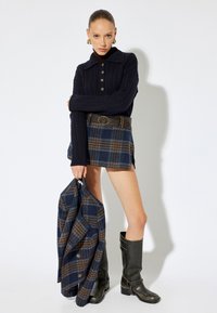 Navy-blauer Kabelstrickpullover mit Kragen und Knöpfen, karierte Rock in Navy und Braun sowie kniehoge schwarze Stiefel. Hält eine passende karierte Jacke.