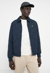 Marineblau gesteppte Jacke mit Reißverschluss und Wellenmuster. Verfügt über einen Kragen und ein gelbes Logo auf der linken Brust. Trägt man über einem weißen Hemd.