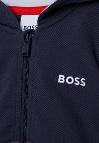 Marinblå zip-up hoodie med strukturerad tyg, röd innervadding och vit broderad "BOSS"-logotyp på bröstet.