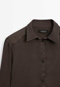 Chemise marron à manches longues avec boutons, col et étiquette de marque visible à l'intérieur du col.