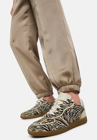 Black and white zebra print sneakers with a textured upper, cream laces, and tan rubber outsole, paired with beige satin joggers.