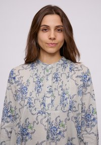 Ljusgrå blus med blå och grön blommönster, plisserad front och rund krage. Tillverkad av mjukt tyg med en lätt texturerad känsla.