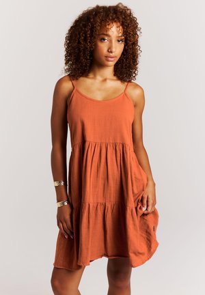 Vrouw met krullend haar draagt een mouwloze oranje jurk, houdt de zoom vast, geaccentueerd met twee gouden armbanden en een neusring, staande tegen een witte achtergrond.