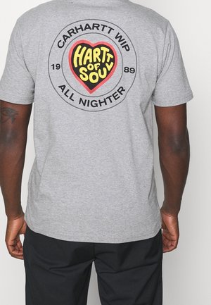 Osoba o ciemnej karnacji skóra, ubrana w szarą koszulkę z okrągłym logo z napisem "Carhartt WIP All Nighter 1989" oraz tekstem w kształcie serca "Hartt of Soul" na plecach.