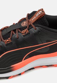 Chaussure de sport noire avec une tige en mesh texturé, des lacets orange distinctifs et une bande orange frappante le long de la semelle.
