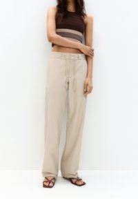Kvinde iført beige bredbenede bukser med snøre i taljen, en ærmeløs brun stribet crop top og brune strop sandaler.