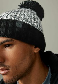 Berretto lavorato a maglia bianco e nero con pom-pom, caratterizzato da un motivo testurizzato e un piccolo etichetta logata nera sulla visiera.